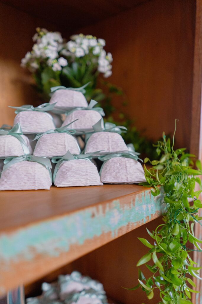 Elegant wedding favor boxes with green ribbon on a rustic shelf, surrounded by greenery.